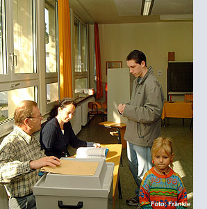 Personen in einem Klassenraum mit Holztischen und Fenstern, zwei Erwachsene und zwei Kinder, helle Wände