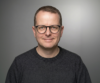 Porträt eines Mannes mit Brille, grauem Pullover aus Strickmaterial, vor grauem Hintergrund