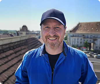Mann mit blauer Jacke und schwarzer Kappe aus Stoff, lächelnd auf Dachterrasse bei Tageslicht