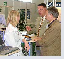 Drei Personen im Gespräch an Messestand mit Metallgestell und Computer, helle Kleidung, braune und beige Töne