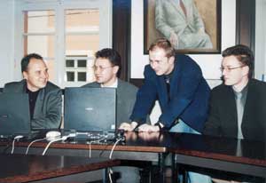 Vier Männer in Anzügen an einem Holztisch mit einem schwarzen Laptop in einem Büro